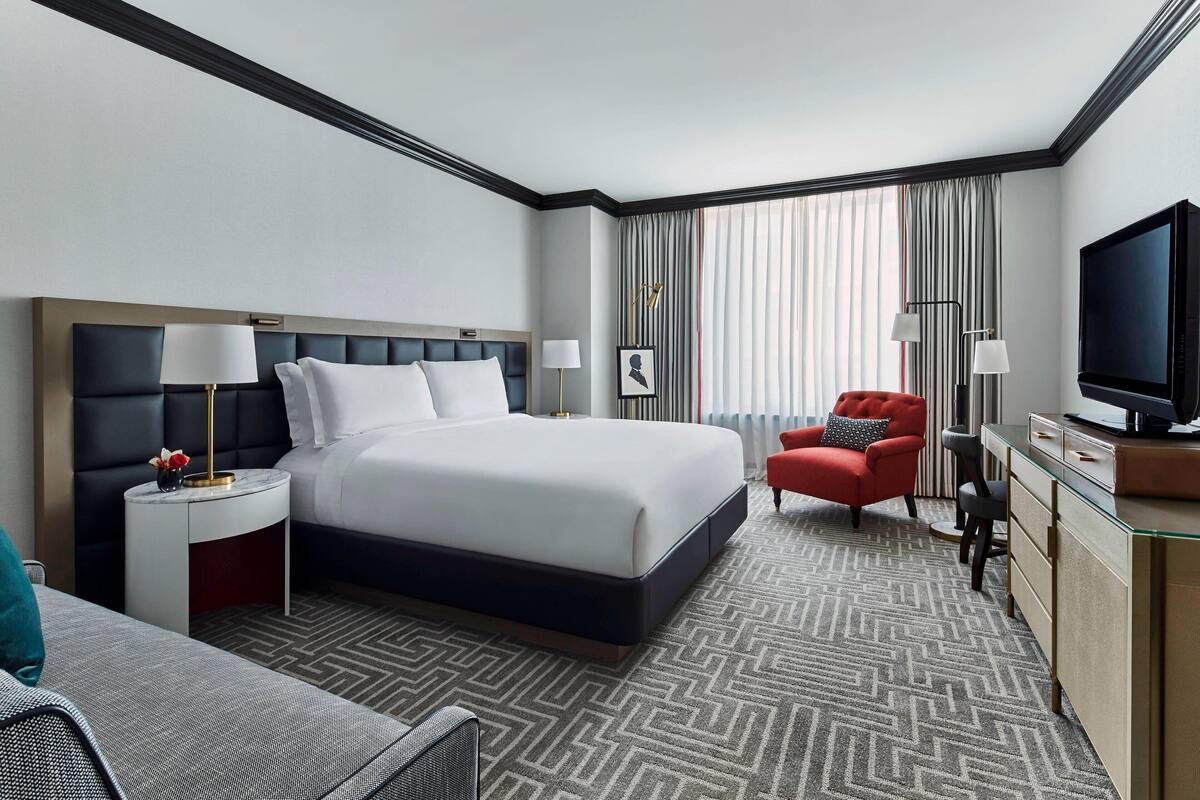 A hotel room with a large bed, a desk with a television, a red armchair, and a nightstand with lamps.