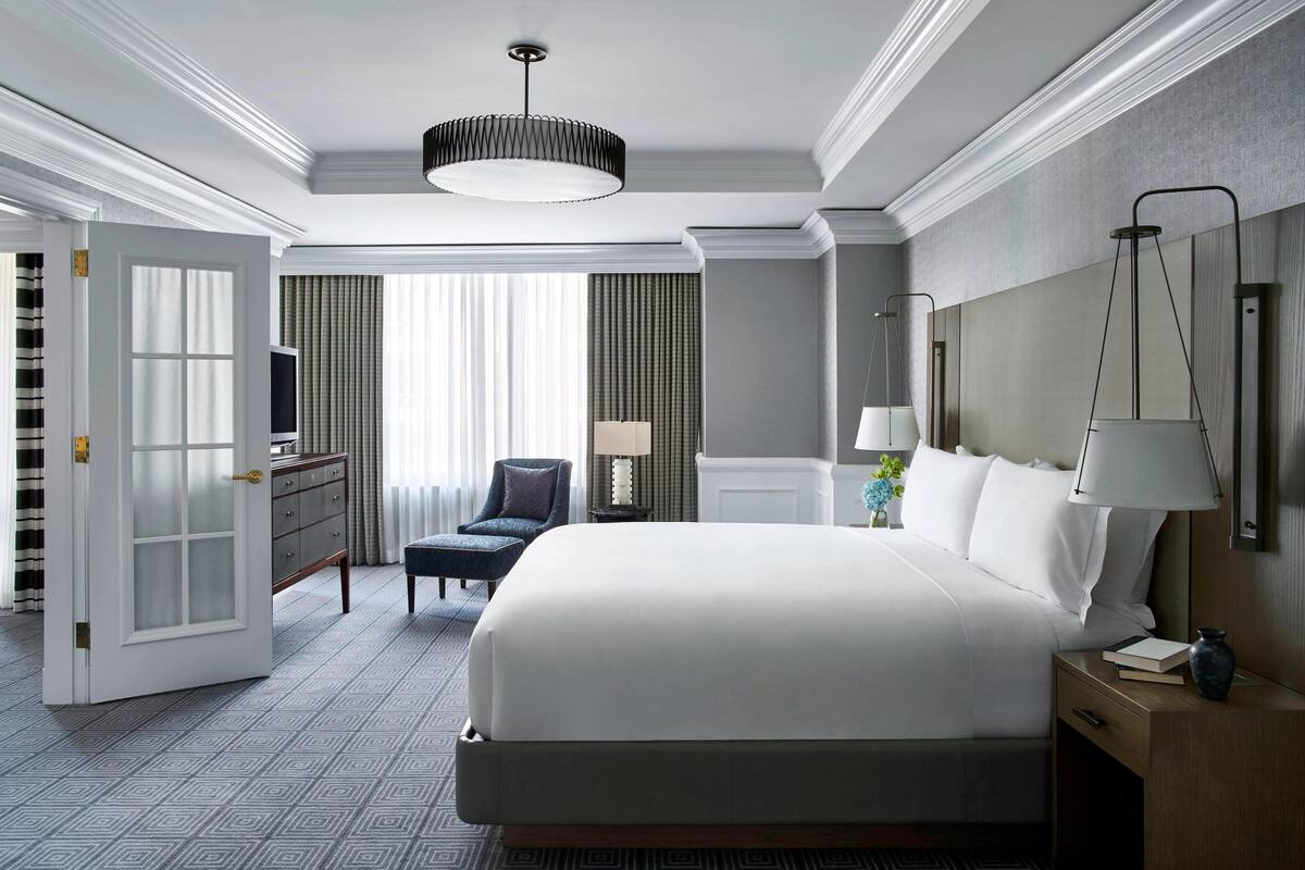 A hotel room with a large bed, a desk, a chair, and a nightstand with a lamp.