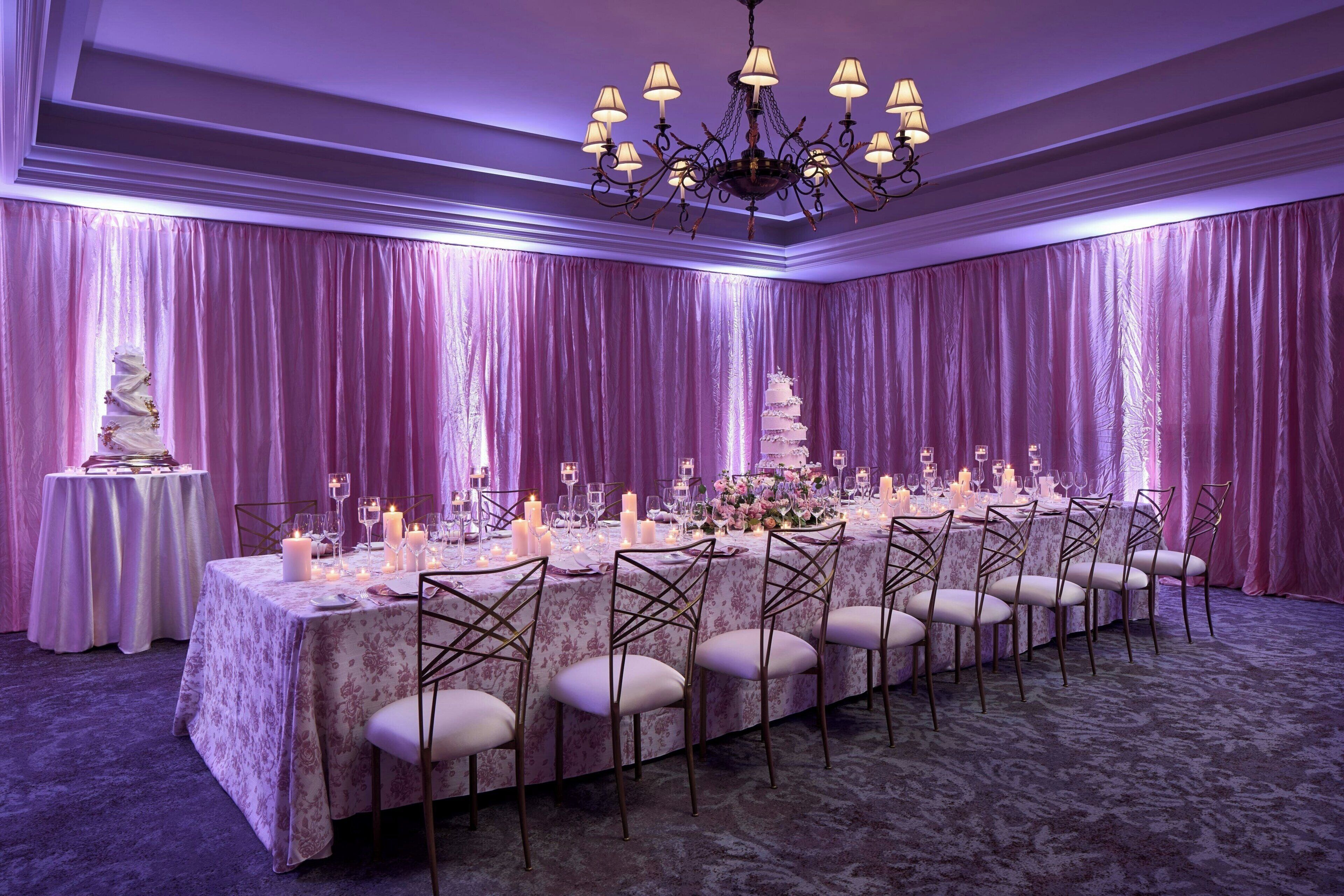 Une salle de banquet avec une longue table dressée pour un événement formel, entourée de rideaux drapés et surmontée d’un lustre.