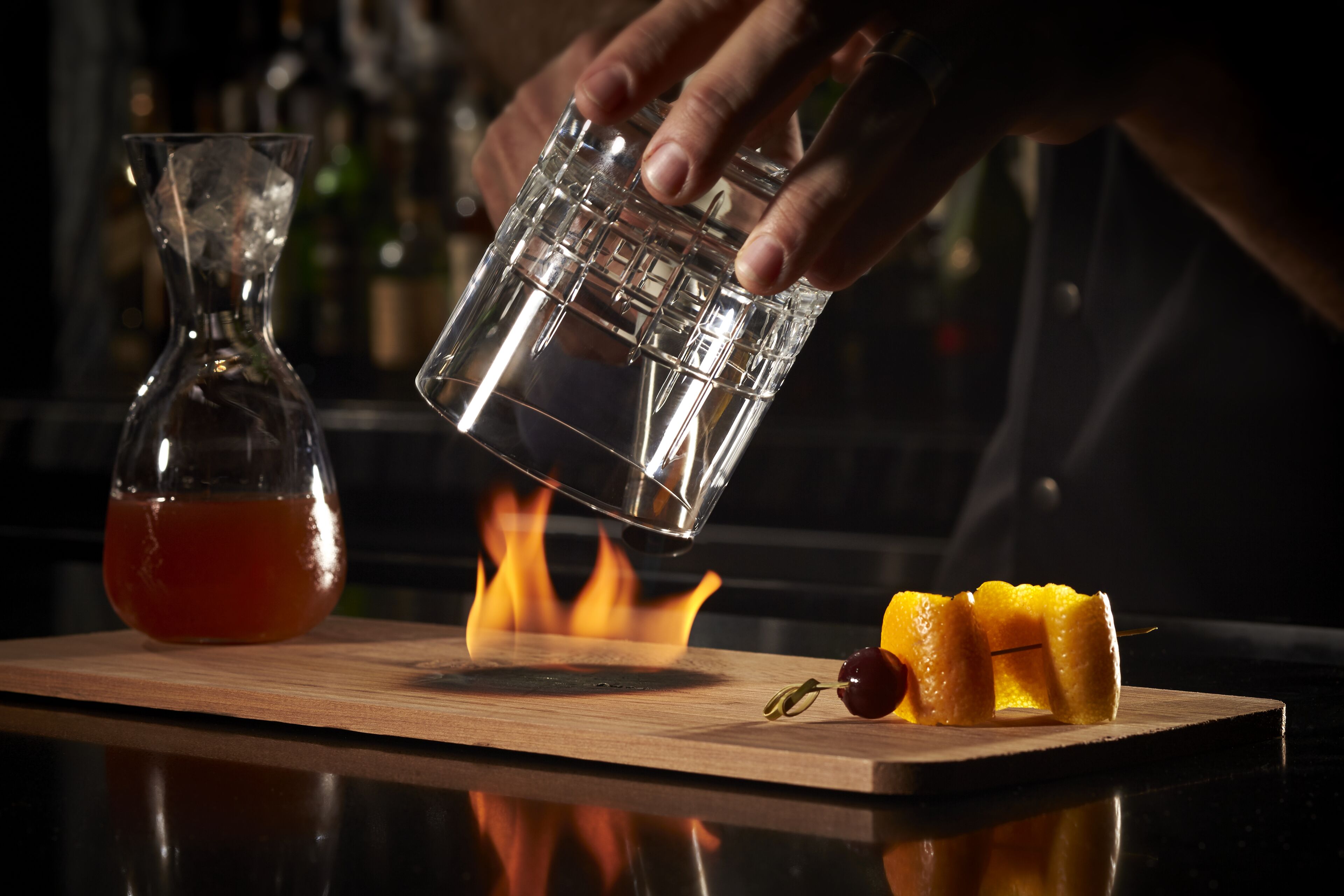 Een hand giet een drankje van een glas in een ander glas op een houten plank, met een fles en fruit.