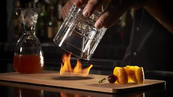 Een hand giet een drankje van een glas in een ander glas op een houten plank, met een fles en fruit.