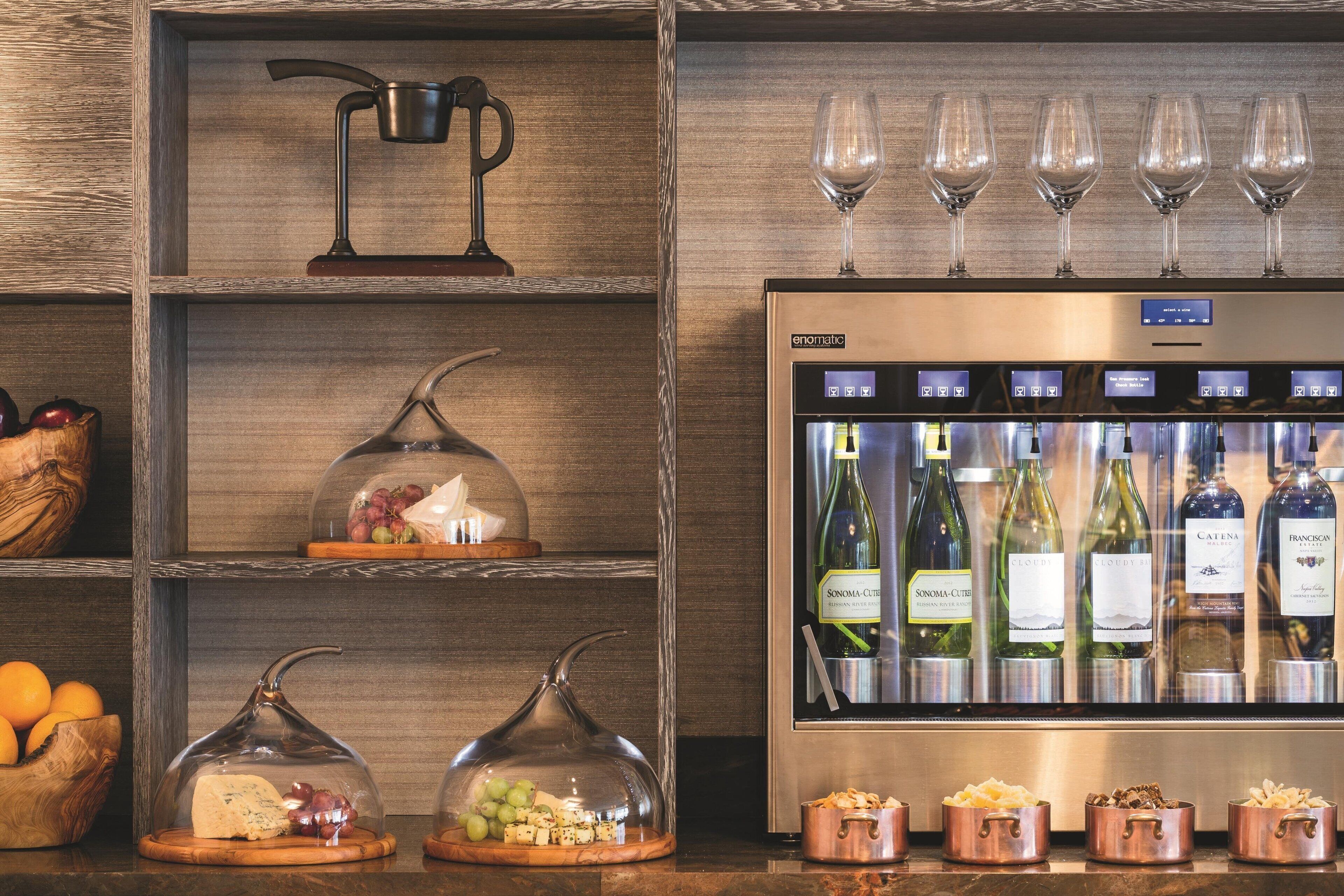 A wine refrigerator stocked with various bottles, a selection of cheeses and grapes on wooden boards, and a wine glass display on the shelf.