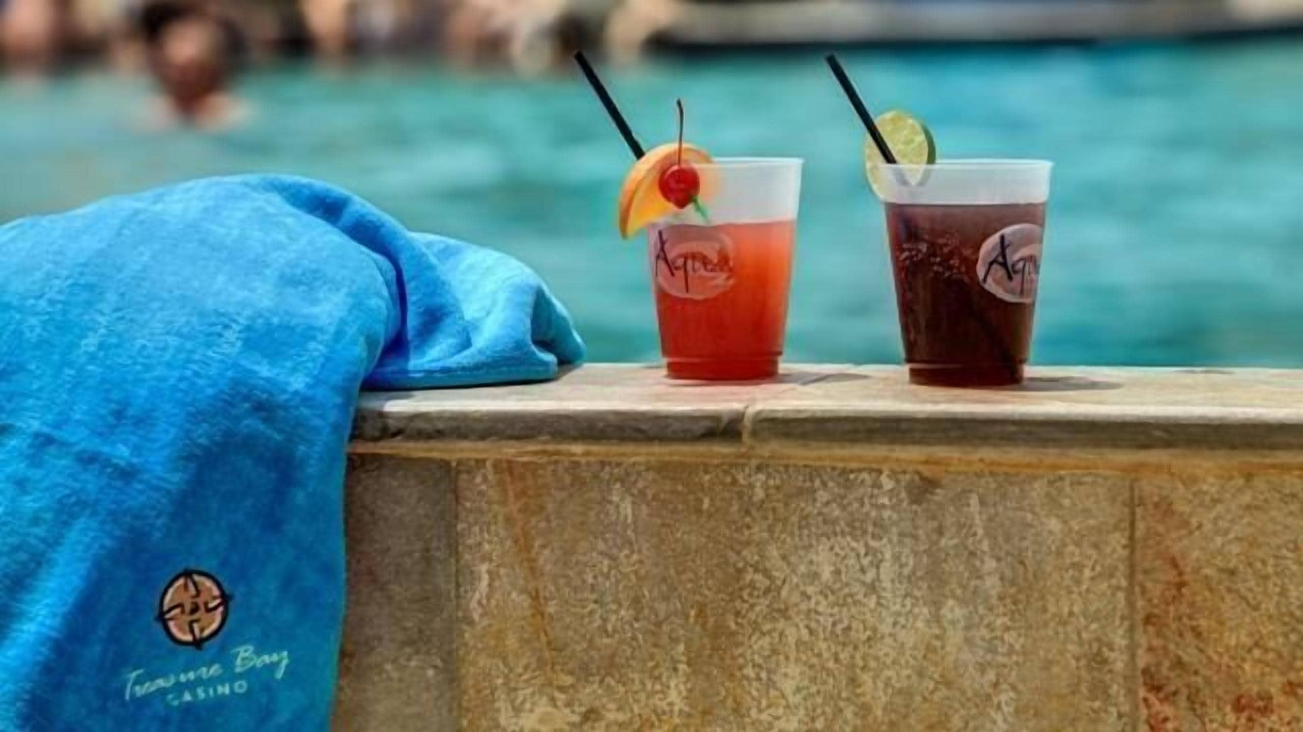 Two drinks with straws and garnishes on a poolside ledge.