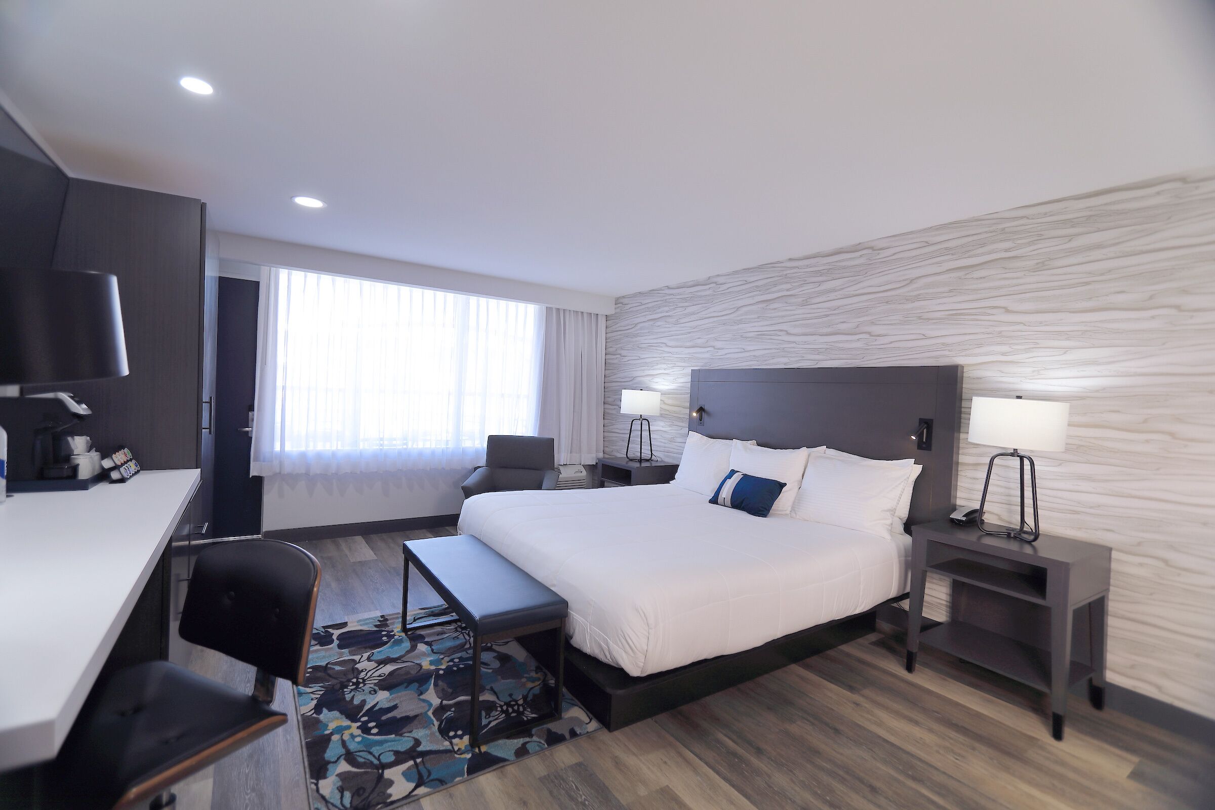 A modern hotel room with a large bed, a desk, a chair, a small table, and a nightstand with a lamp.