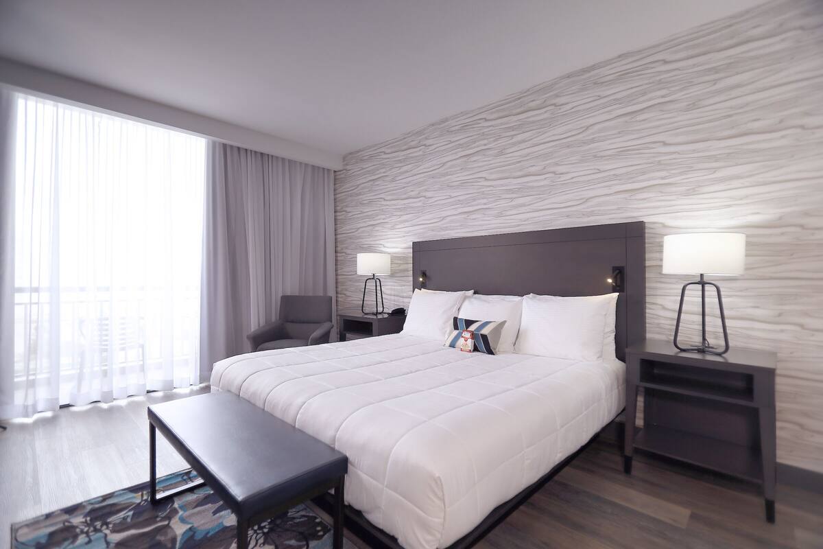 A modern hotel room with a large bed, a desk, a chair, and a view of the city through large windows.