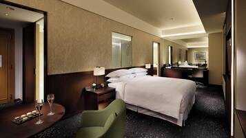 A hotel room with a bed, a desk with a chair, a mirror, and a small table with two glasses of wine.