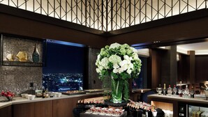 A modern hotel lobby with a checkered floor, a central bar area with a floral arrangement, and a city view through large windows.