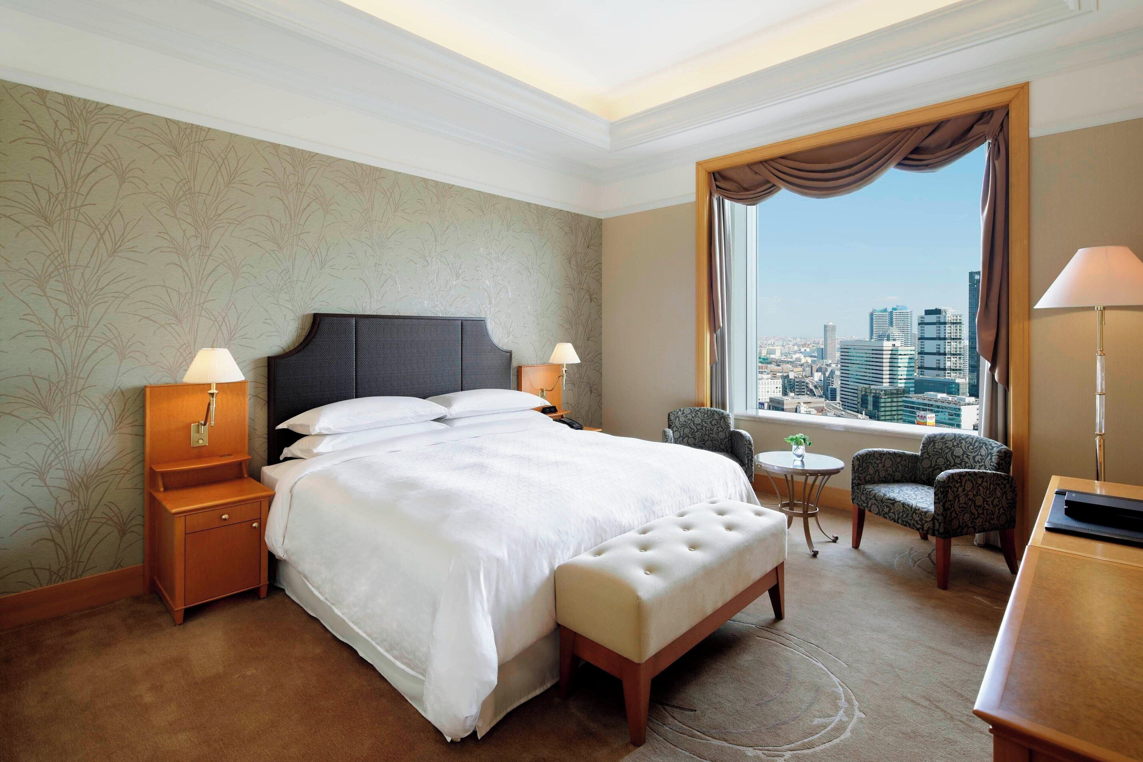 Une chambre d’hôtel avec un grand lit, deux tables de chevet avec des lampes, une petite table ronde avec une chaise, et une vue sur la ville par une grande fenêtre.
