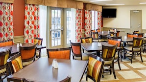 A hotel dining area with multiple tables and chairs, large windows with patterned curtains, and a flat-screen TV mounted on the wall. - Quality Inn Vicksburg (Vicksburg)
