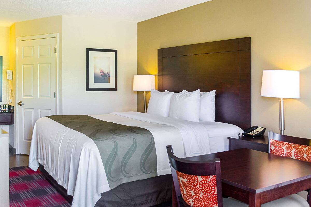 A hotel room with a bed, bedside table, lamp, chair, and a desk with a telephone.