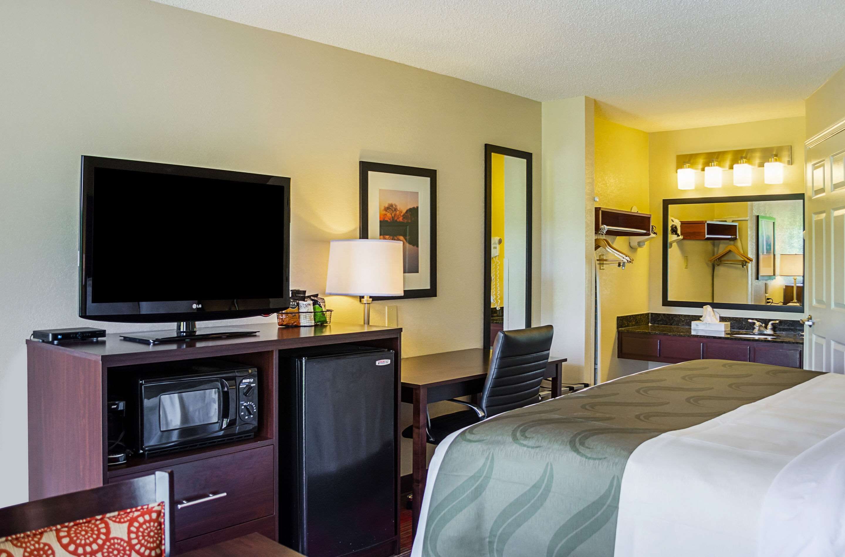 Une chambre d’hôtel comprenant un lit, un bureau avec une télévision, un four à micro-ondes, un réfrigérateur, une chaise et une salle de bain avec un miroir et un lavabo.