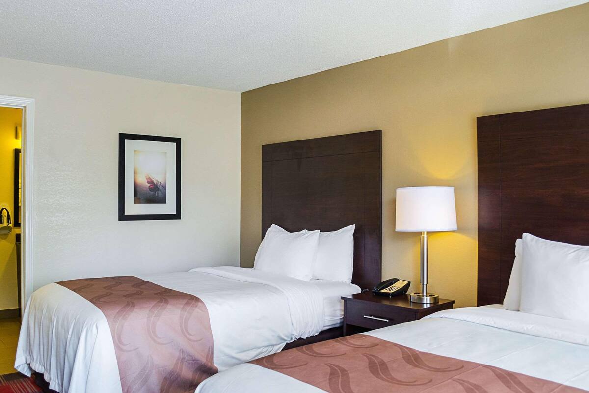 A hotel room with two beds, a bedside table with a lamp, a framed picture on the wall, and a door to the left.