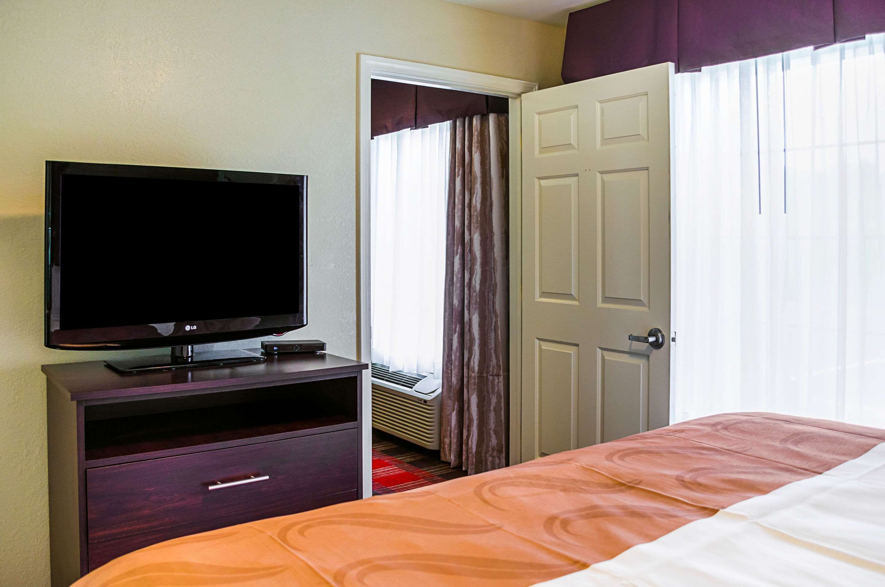 A hotel room with a bed, a TV on a wooden stand, a door, and a window with curtains.