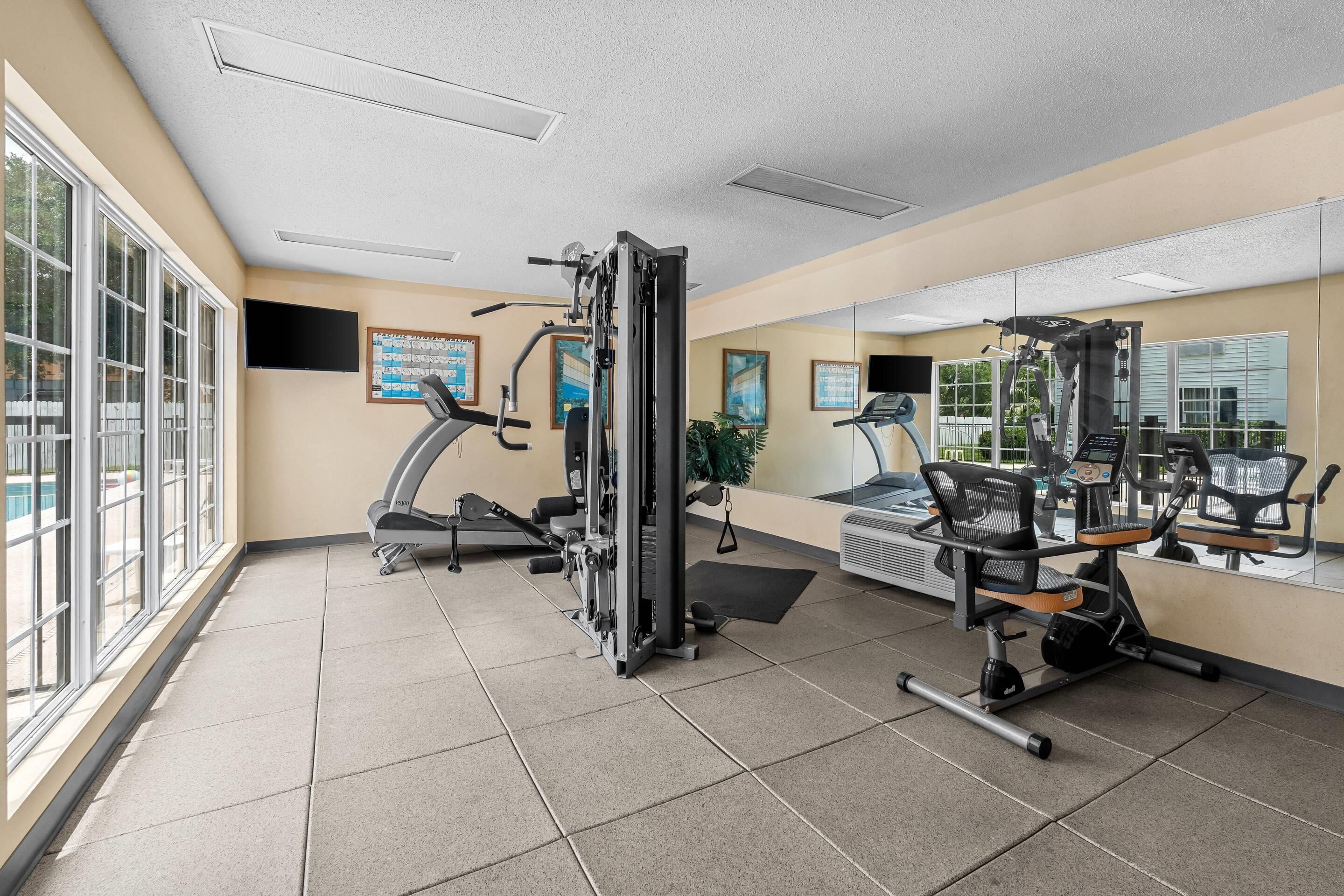 Un gimnasio con equipamiento de ejercicio, ventanas amplias y un muro de espejos.