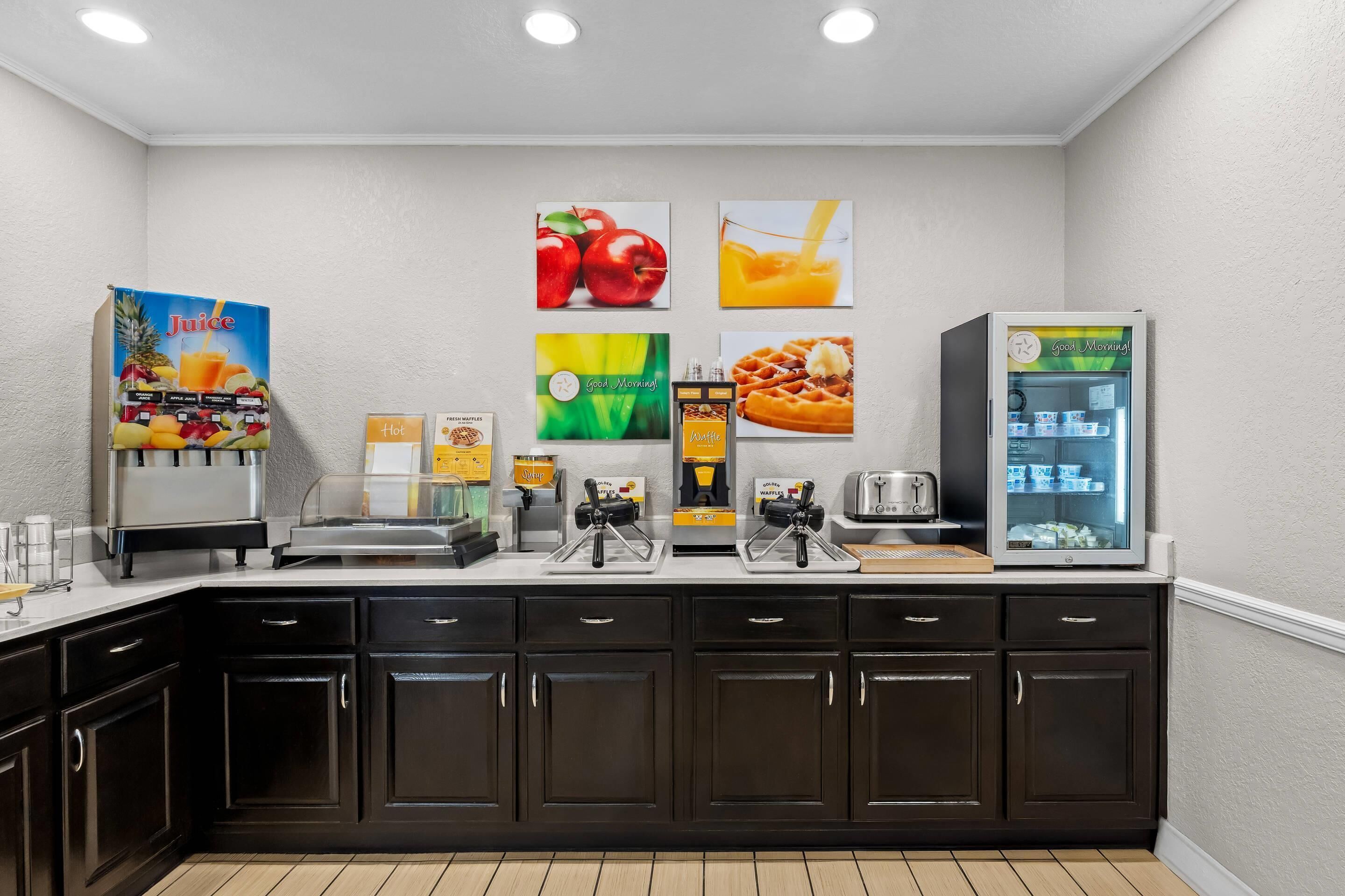 A breakfast bar with juice dispensers, waffle maker, toaster, and a refrigerator.