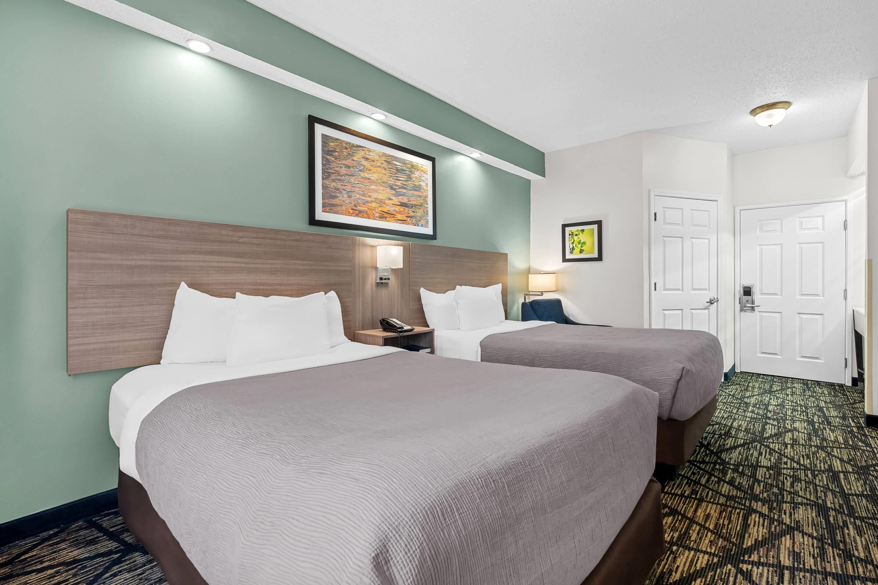 A hotel room with two beds, a headboard, wall-mounted lamps, a framed artwork, and a carpeted floor.