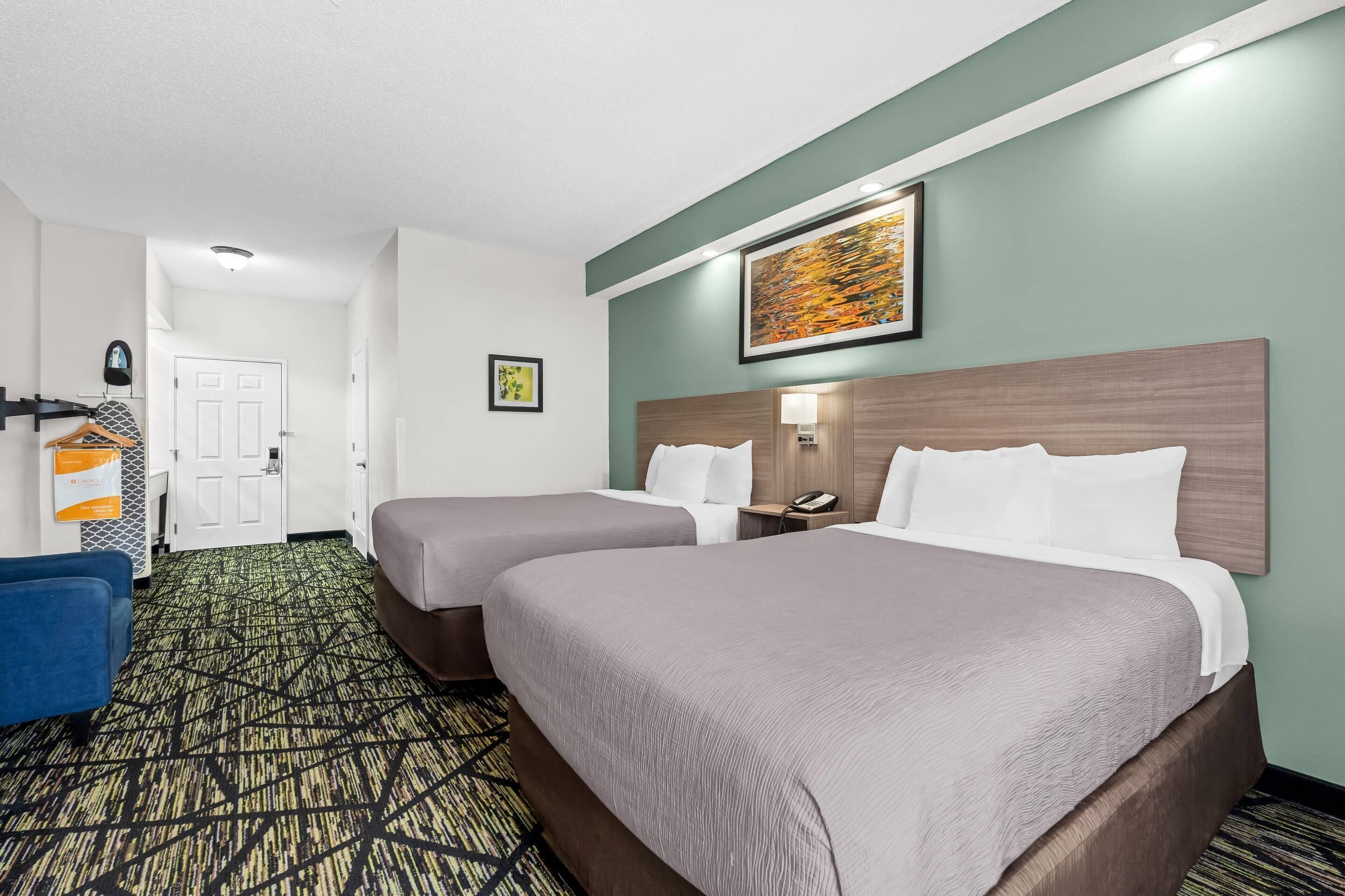 A hotel room with two beds, a blue chair, a patterned carpet, a wooden headboard, a wall-mounted phone, and a framed picture.