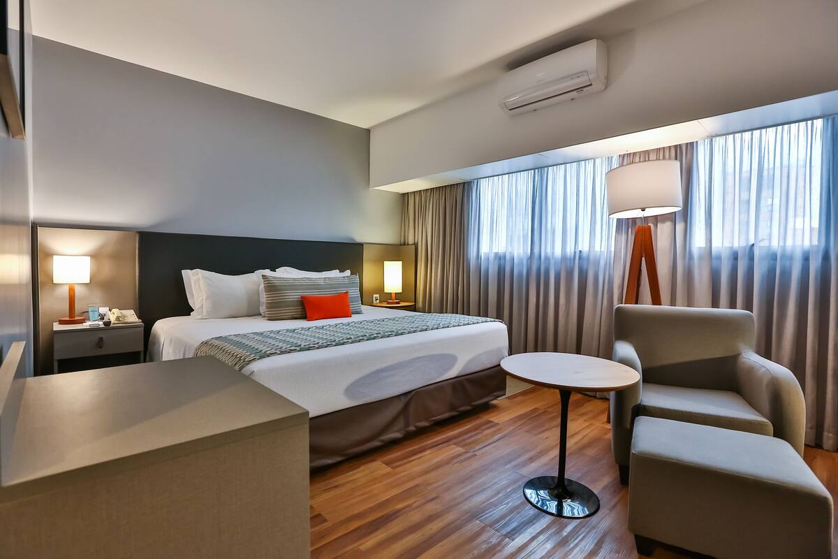 A hotel room with a bed, bedside table, lamp, chair, and a small table.