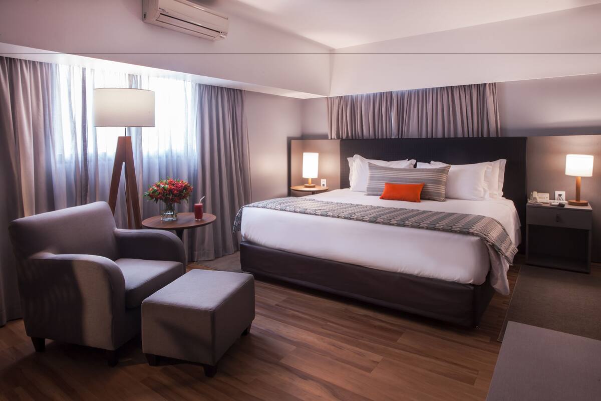 A hotel room with a large bed, a chair, a small table with a vase of flowers, and a lamp.
