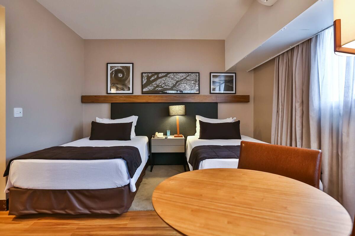 A hotel room with two beds, a small table, and a nightstand with a lamp.