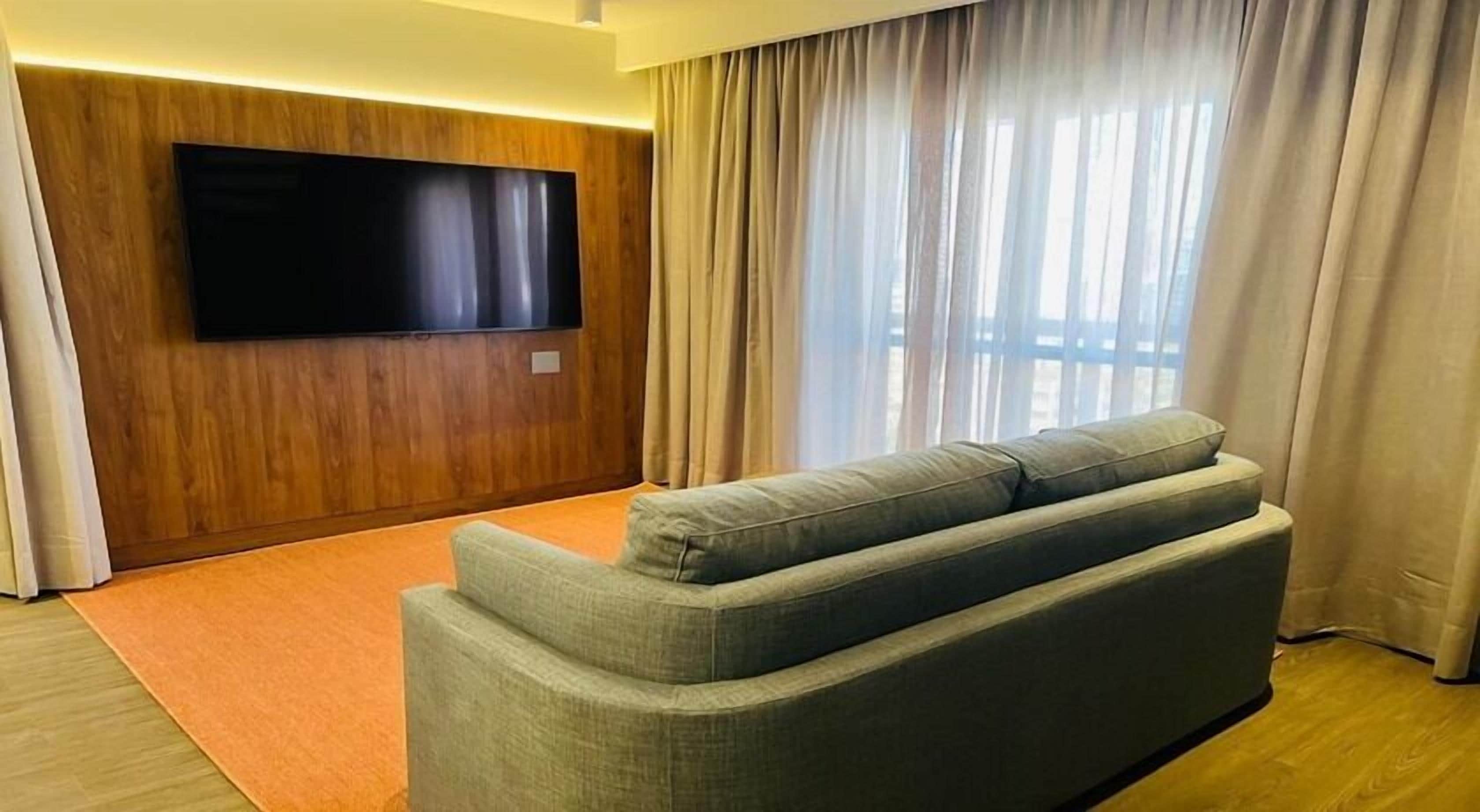 Una sala de estar moderna con un televisor de pantalla plana montado en una pared de madera, un sofá gris y un ventanal con cortinas.