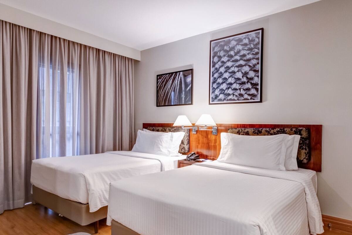 Ein Hotelzimmer mit zwei Betten, einem großen Fenster mit Vorhängen und zwei gerahmten Kunstwerken an der Wand.
