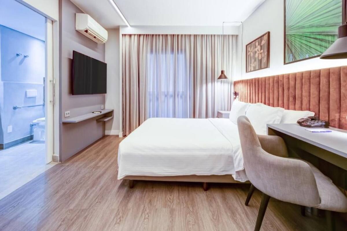 Ein Hotelzimmer mit Bett, Schreibtisch, Sessel, Fernseher und einem fensterbepflanzten Fenster.