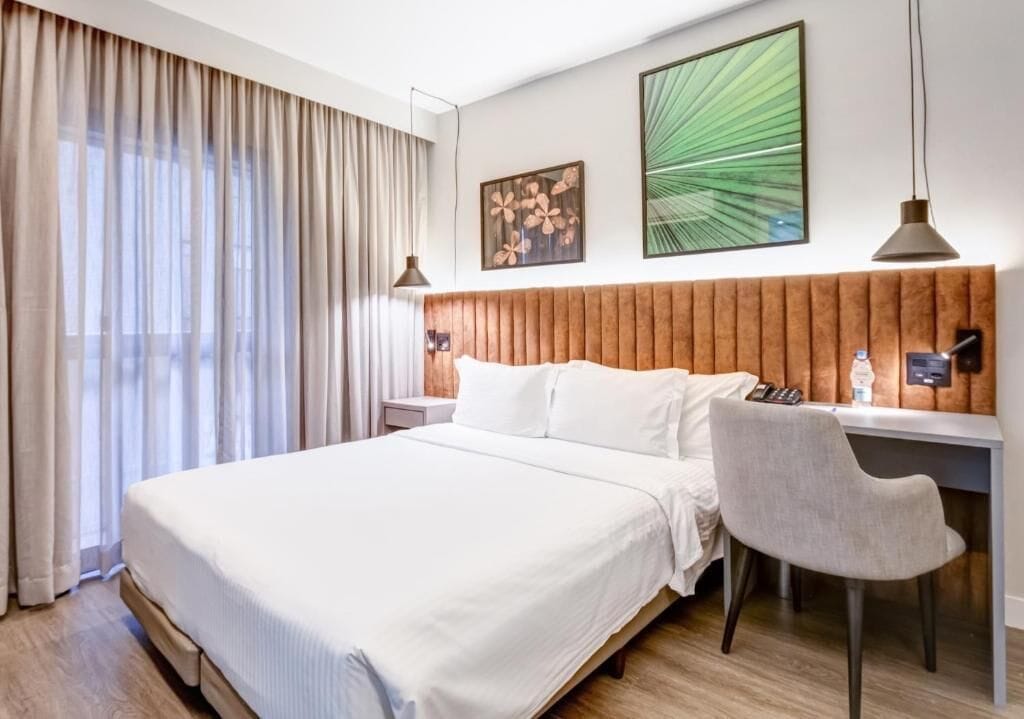 Habitación de hotel con una cama grande, un escritorio y una silla. Tiene una ventana con cortinas, una lámpara de pared y un cuadro enmarcado.