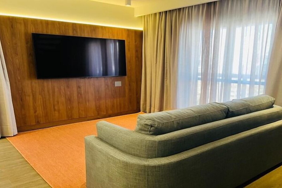 Una sala de estar moderna con un televisor de pantalla plana montado en una pared de madera, un sofá gris y un ventanal con cortinas.