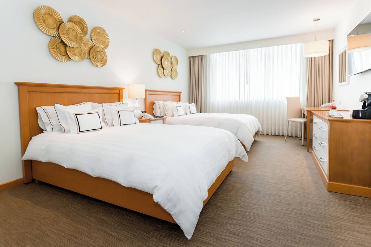 Habitación de hotel con dos camas, cabecero de madera, escritorio con silla, lámpara y una ventana grande con cortinas.