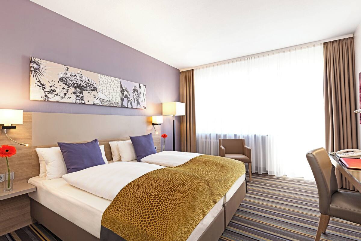 Hotelkamer met een groot bed, twee nachtkastjes, een bureau en een stoel. Er is een raam met gordijnen, een schilderij aan de muur en een lamp op elk nachtkastje.