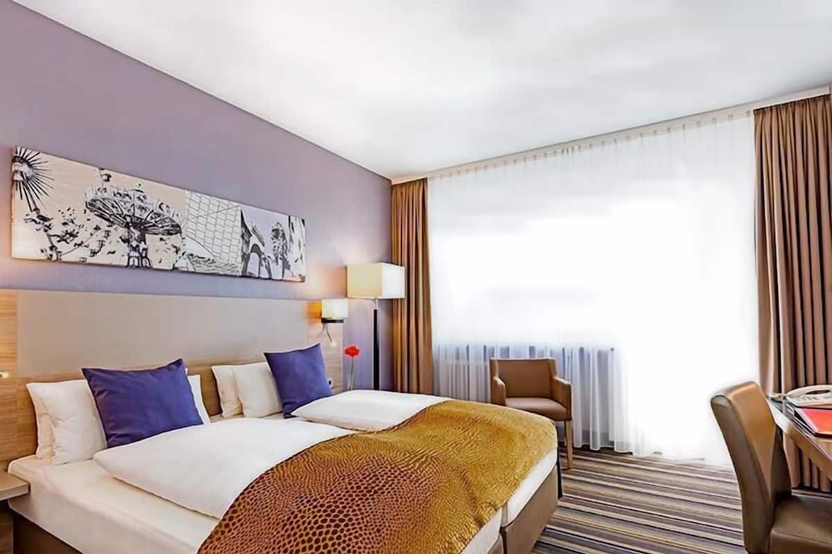 Een hotelkamer met een bed, twee kussens, een nachtkastje, een lamp, een stoel en een raam met gordijnen.