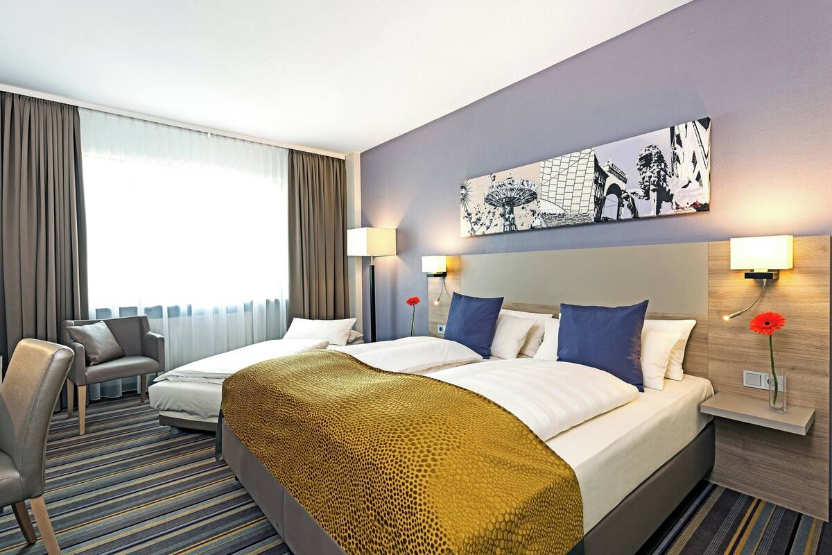 Quarto de hotel com duas camas, um sofá, uma mesa e uma janela grande com cortinas.