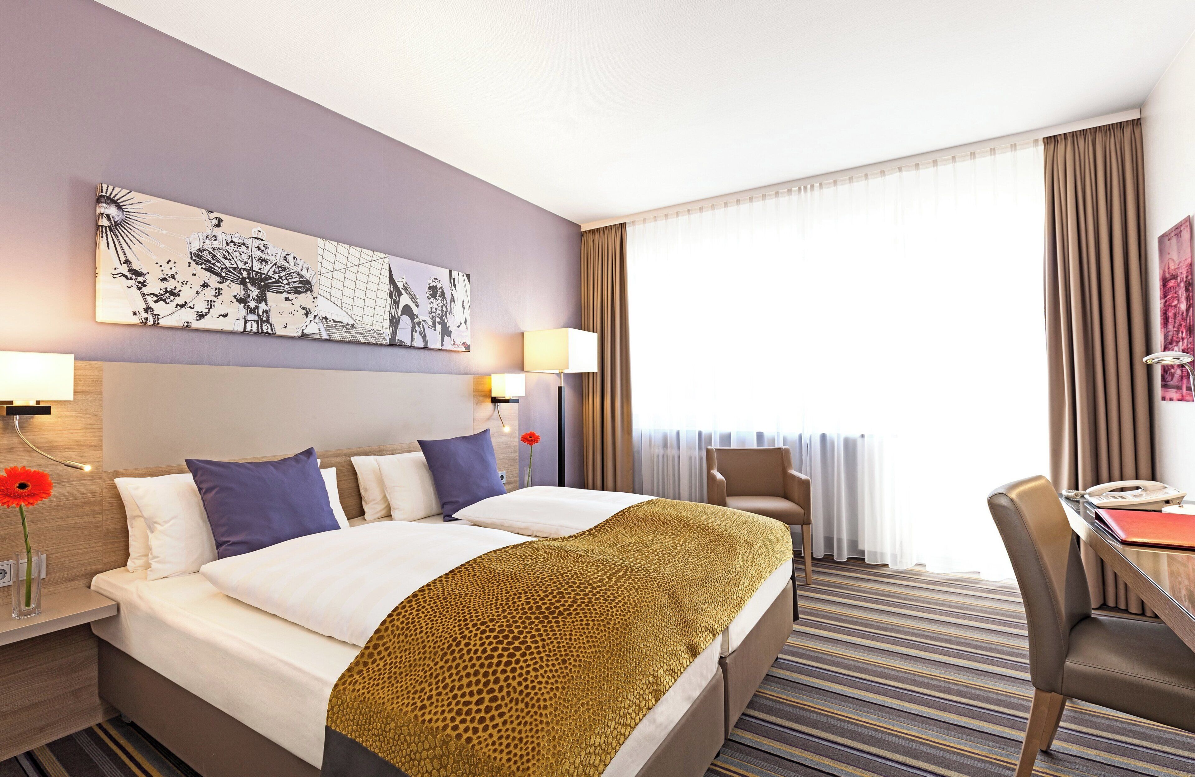 Hotelkamer met een groot bed, twee nachtkastjes, een bureau en een stoel. Er is een raam met gordijnen, een schilderij aan de muur en een lamp op elk nachtkastje.