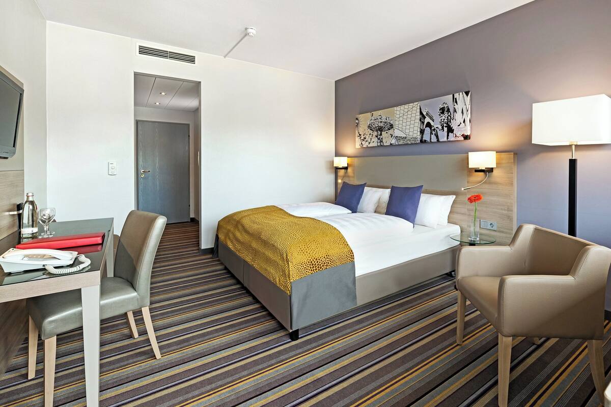 Quarto de hotel com uma cama, uma secretária com telefone, uma cadeira, um abajur e um quadro na parede.