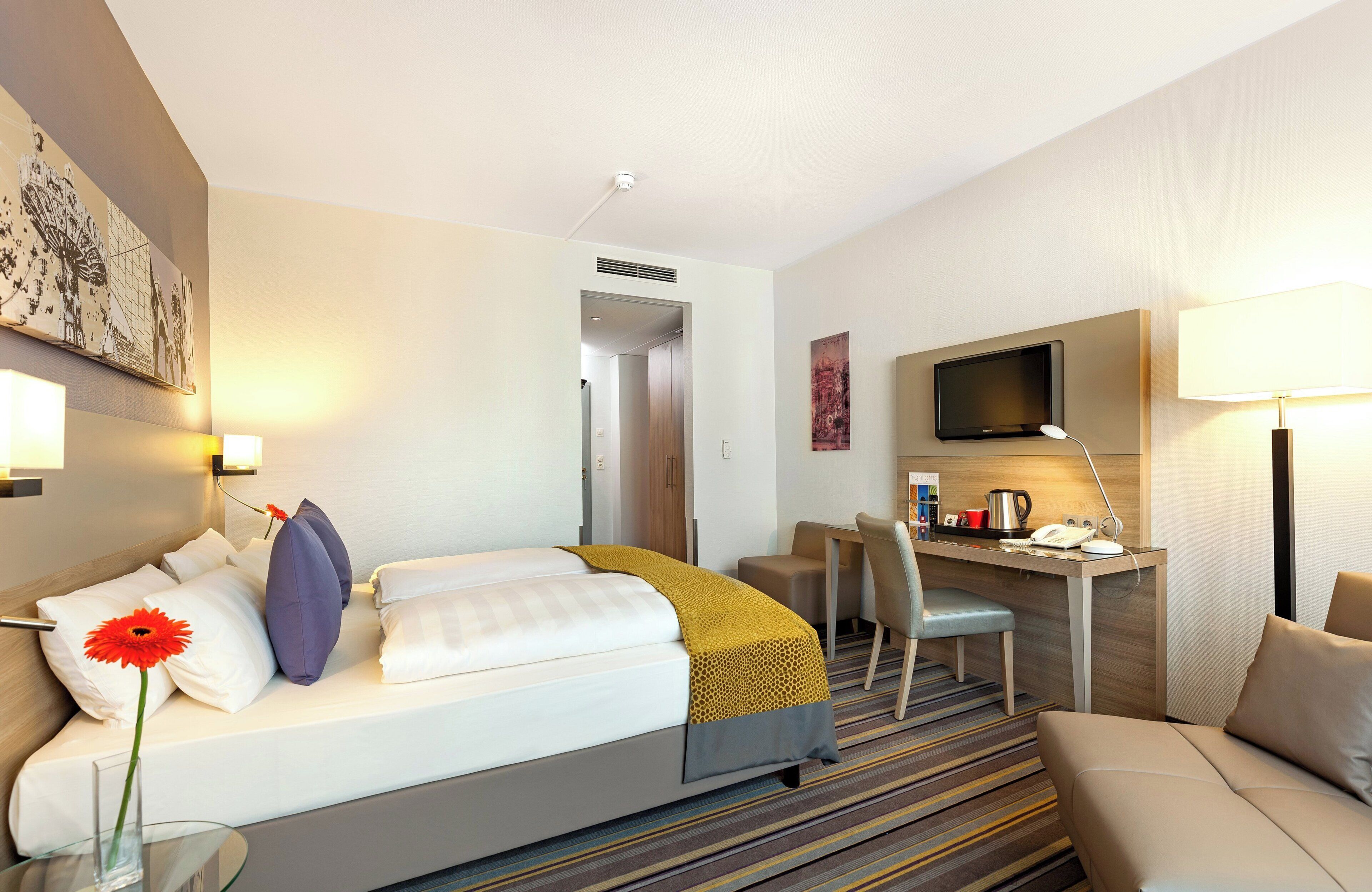 A hotel room with a bed, a desk, a chair, a TV, and a lamp.