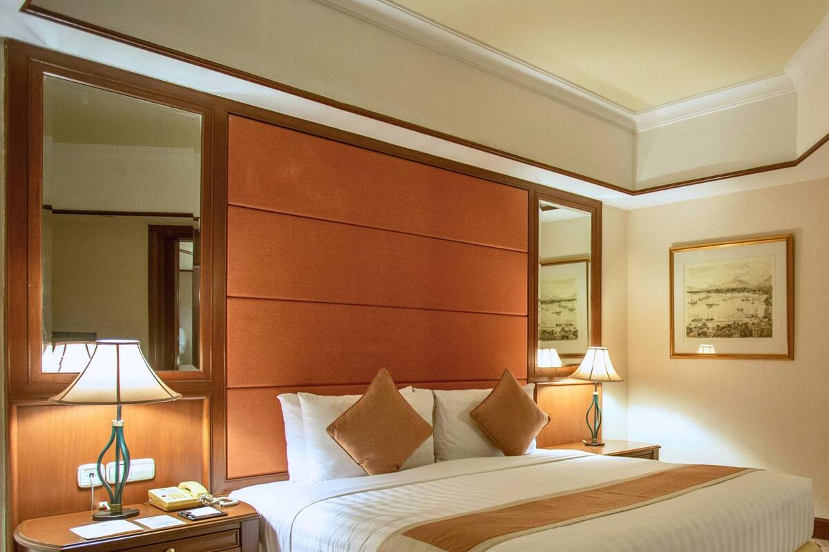 A hotel room with a large bed, two bedside tables with lamps, a mirror, a red armchair, and a small table with a lamp.