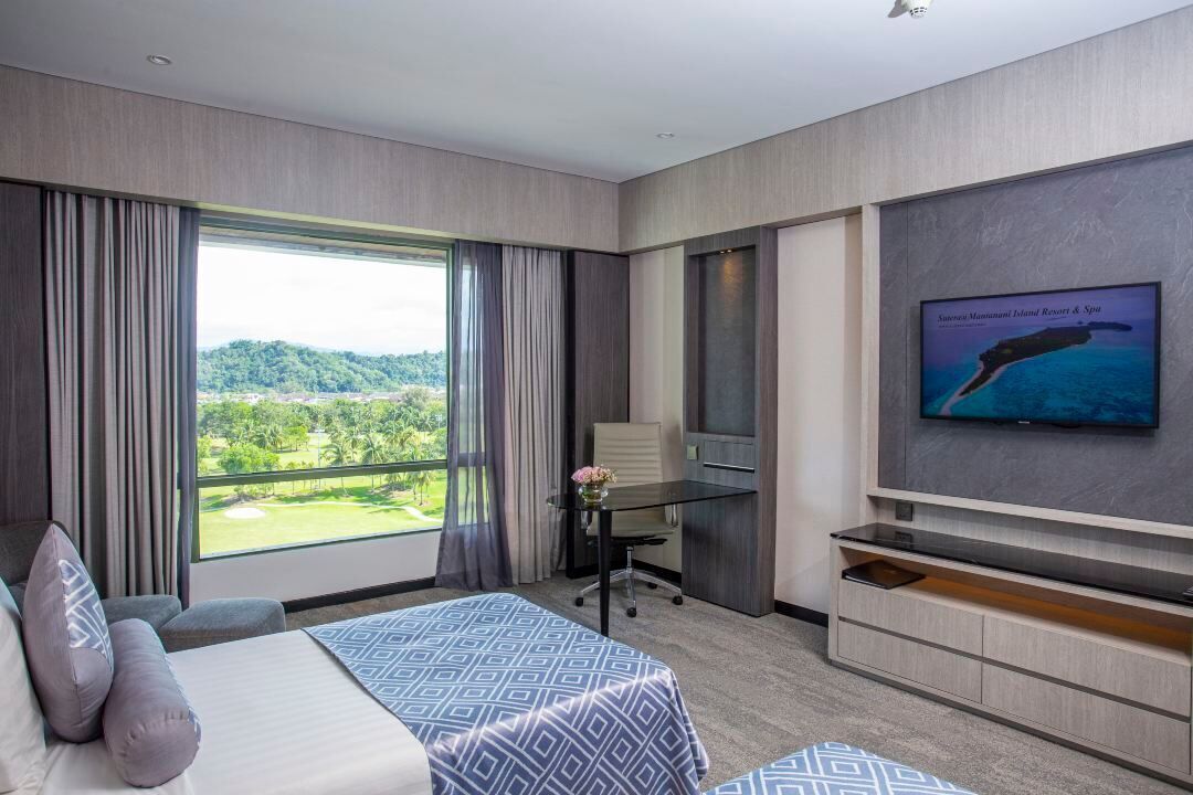 A hotel room with a large window offering a view of a golf course and greenery, a bed with patterned bedding, a desk with a chair, and a flat-screen TV mounted on the wall.