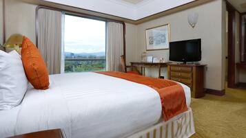 A hotel room with a large bed, a desk with a television, and a window offering a city view.