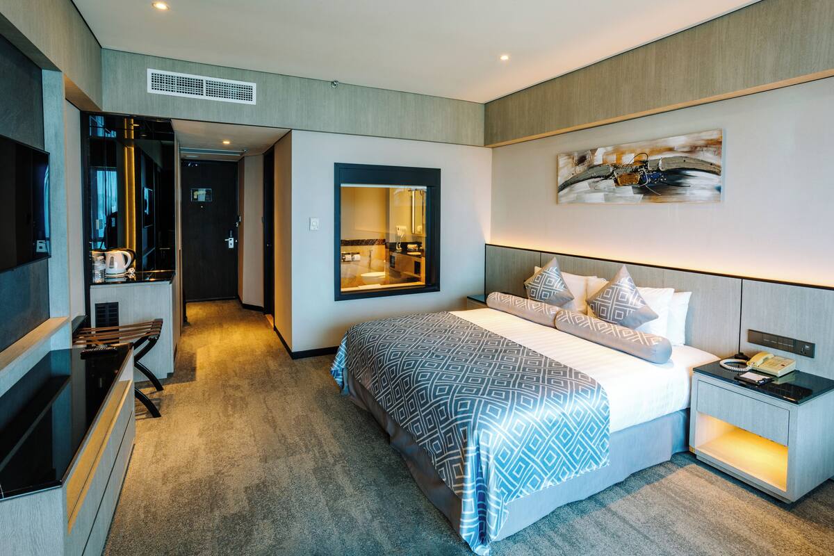 A modern hotel room with a large bed, a bedside table with a lamp, a TV, and a bathroom visible through an open door.