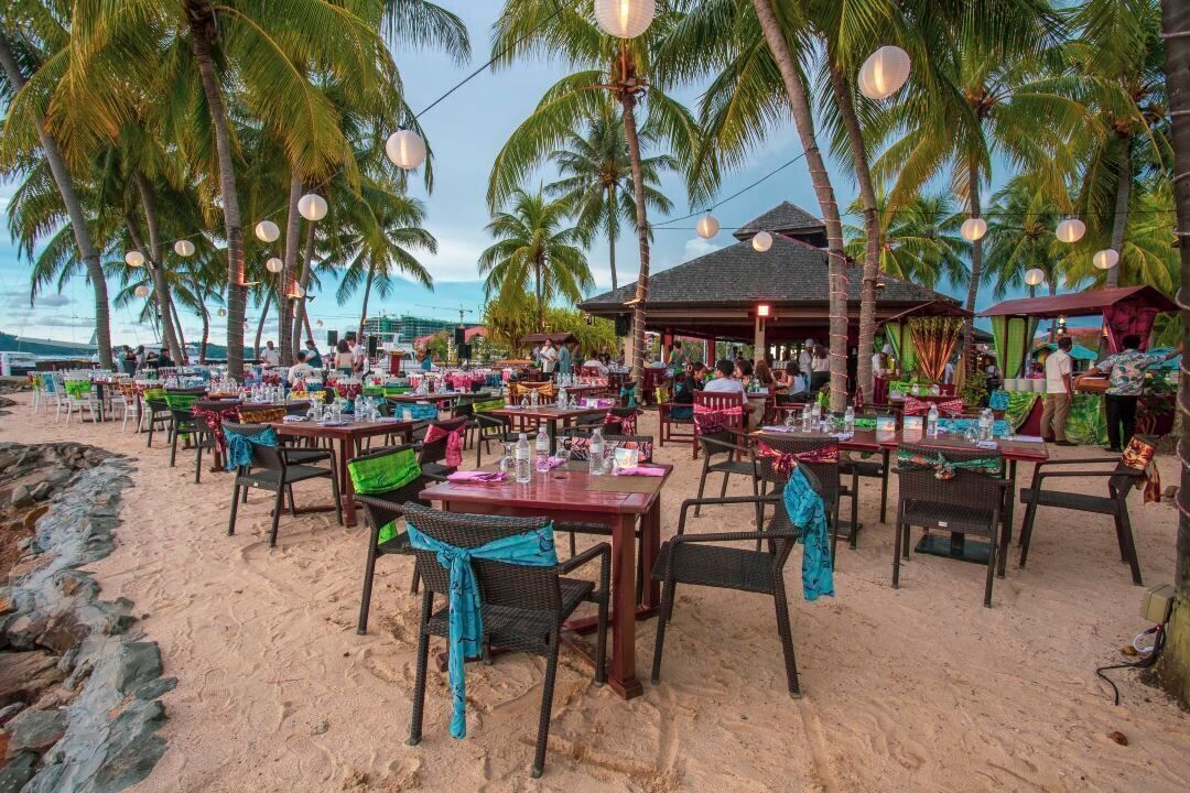 An outdoor dining area with tables and chairs, surrounded by palm trees and a beach view.