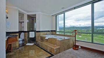 A modern bathroom with a large bathtub, a toilet, and a shower. It features a large window with a view of a green landscape.