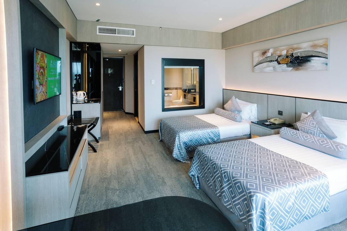 A hotel room with two beds, a TV, a desk, and a bathroom visible through a glass door.
