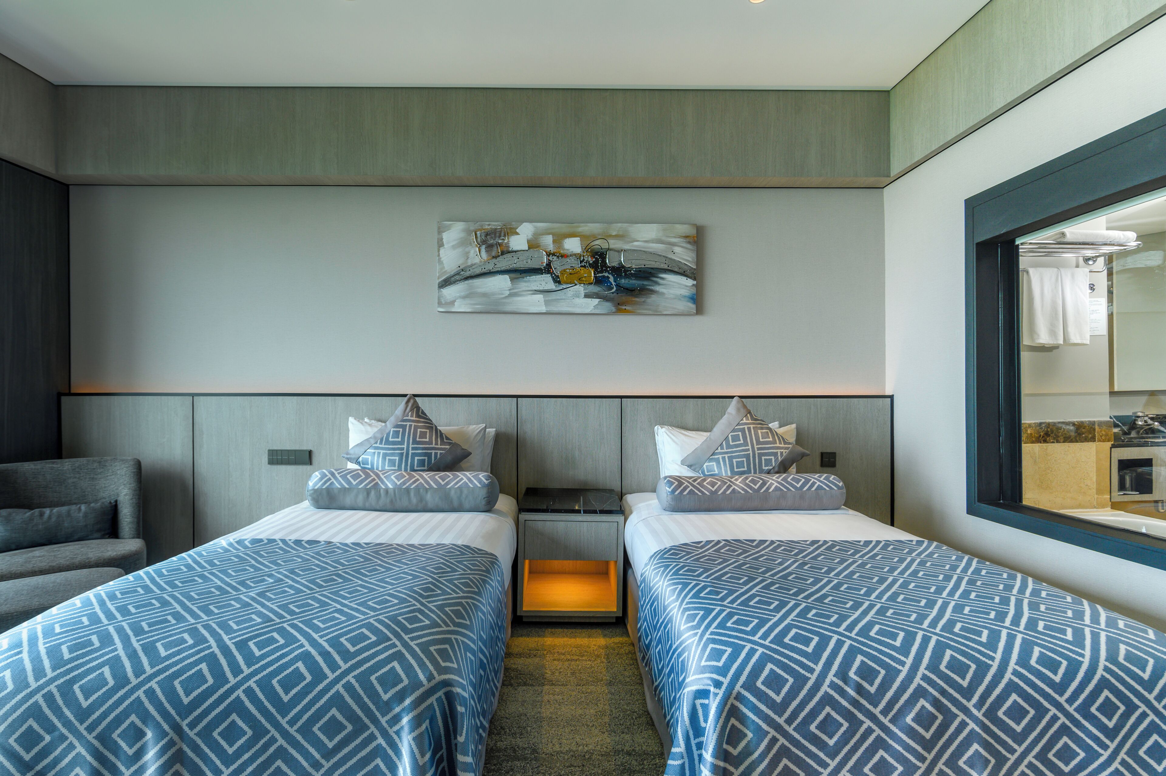 A modern hotel room with two beds, a nightstand, a painting, and a large window with a view of the bathroom.