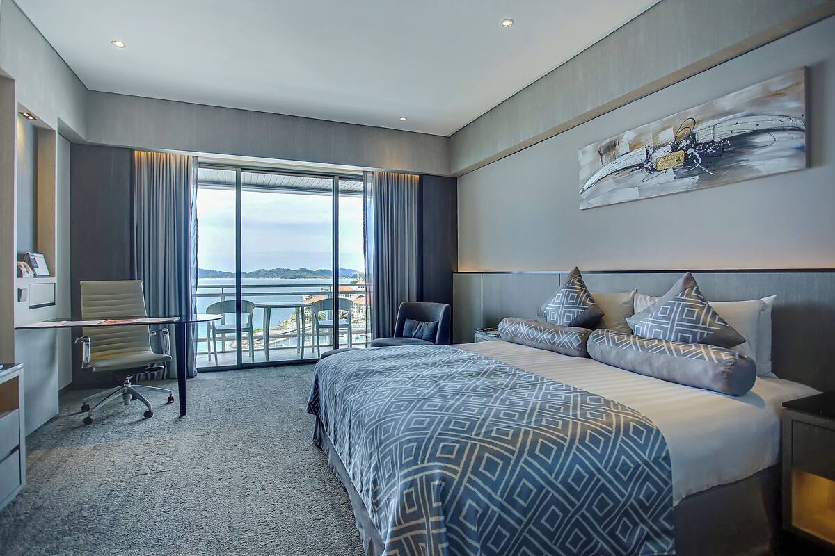 A modern hotel room with a large bed, a desk, and a chair. There is a large window with curtains, a painting on the wall, and a view of the outside.
