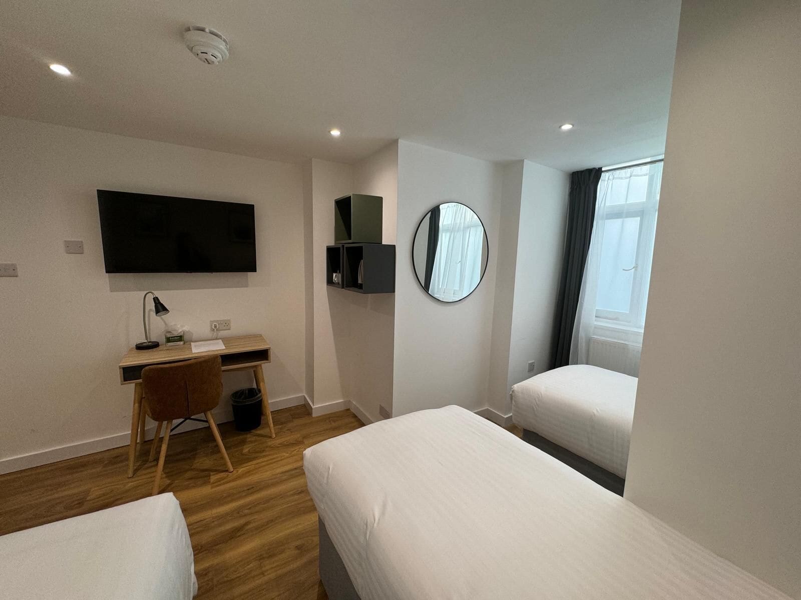 Quarto de hotel com cama, escrivaninha, cadeira, televisão, espelho e janela.