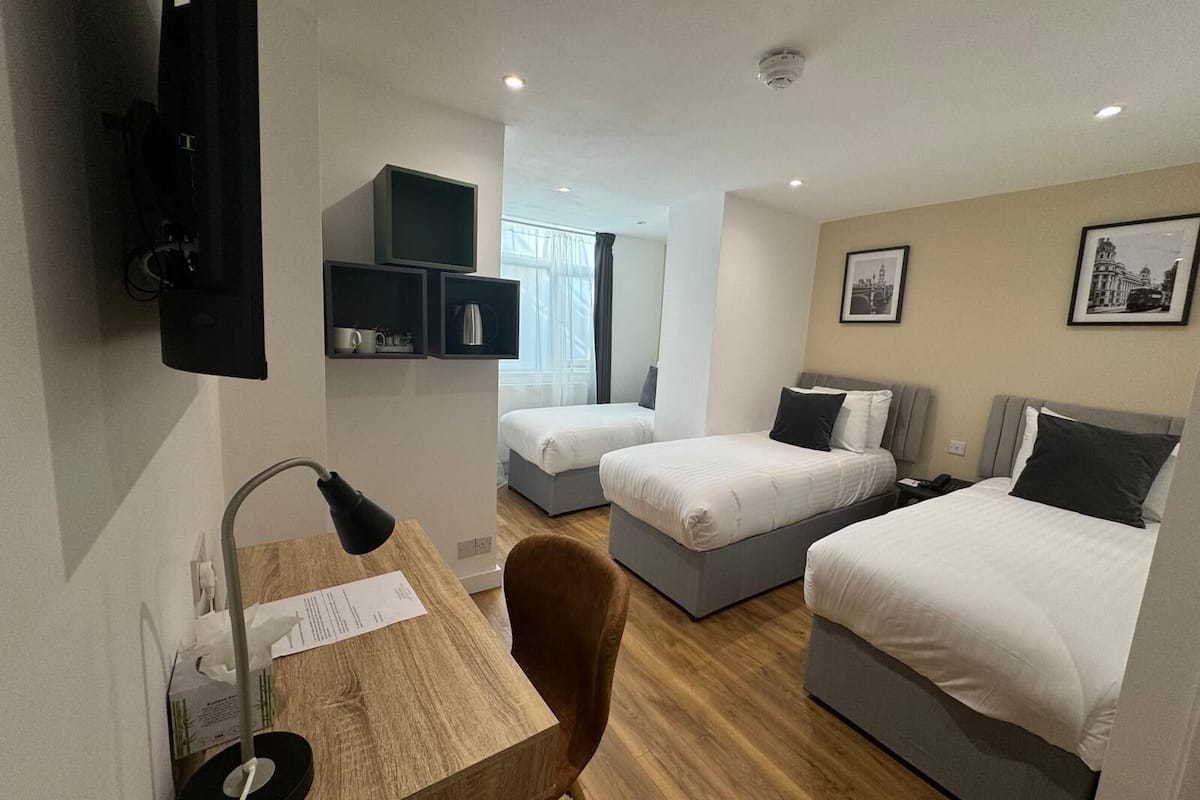 Habitación de hotel con dos camas, un escritorio con lámpara, una silla y una pequeña mesa con un menú. Hay estantes sobre el escritorio y una ventana con cortinas.