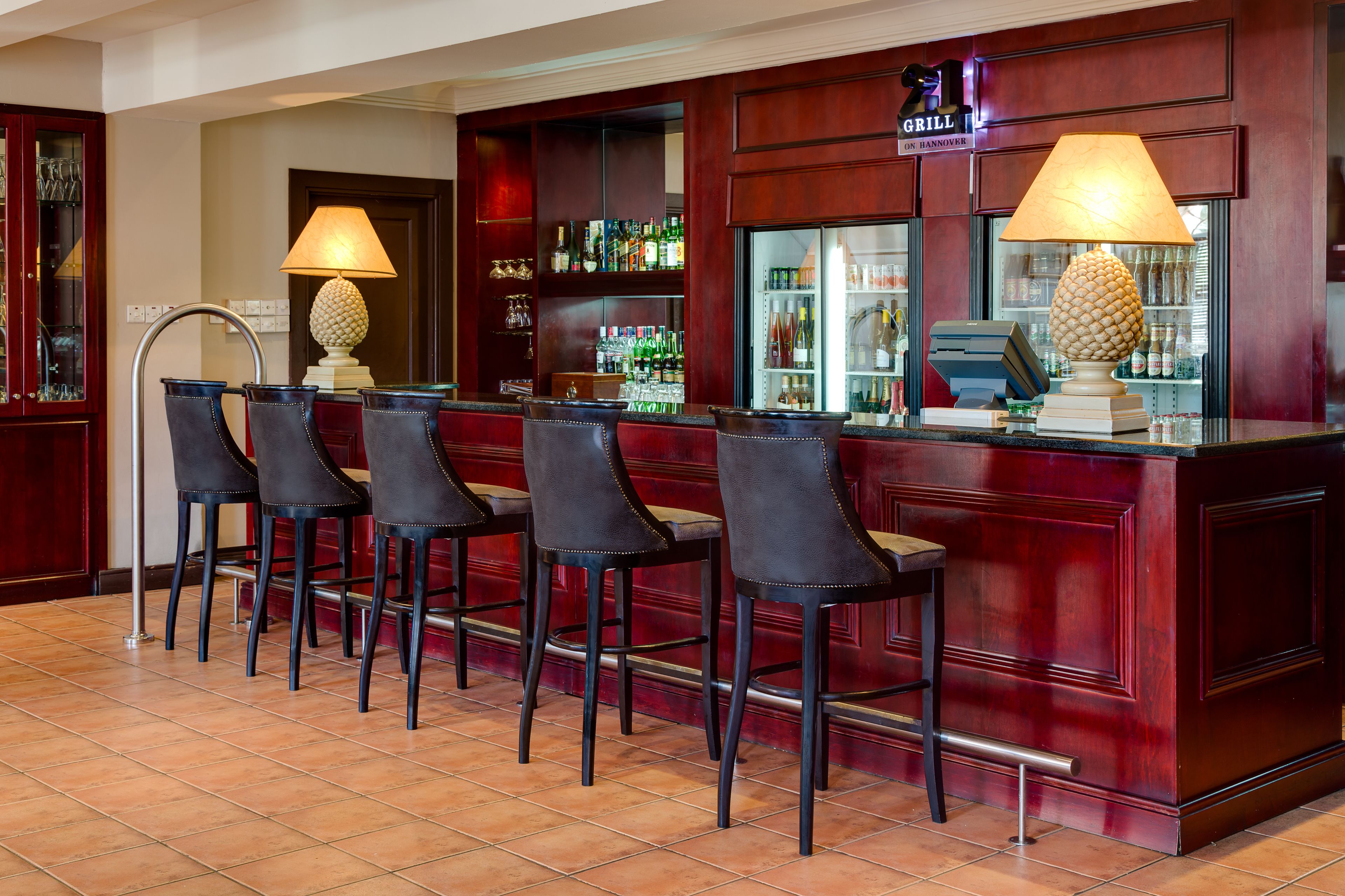 Zona de bar con cuatro taburetes altos, una caja registradora y una variedad de botellas y vasos en estanterías.