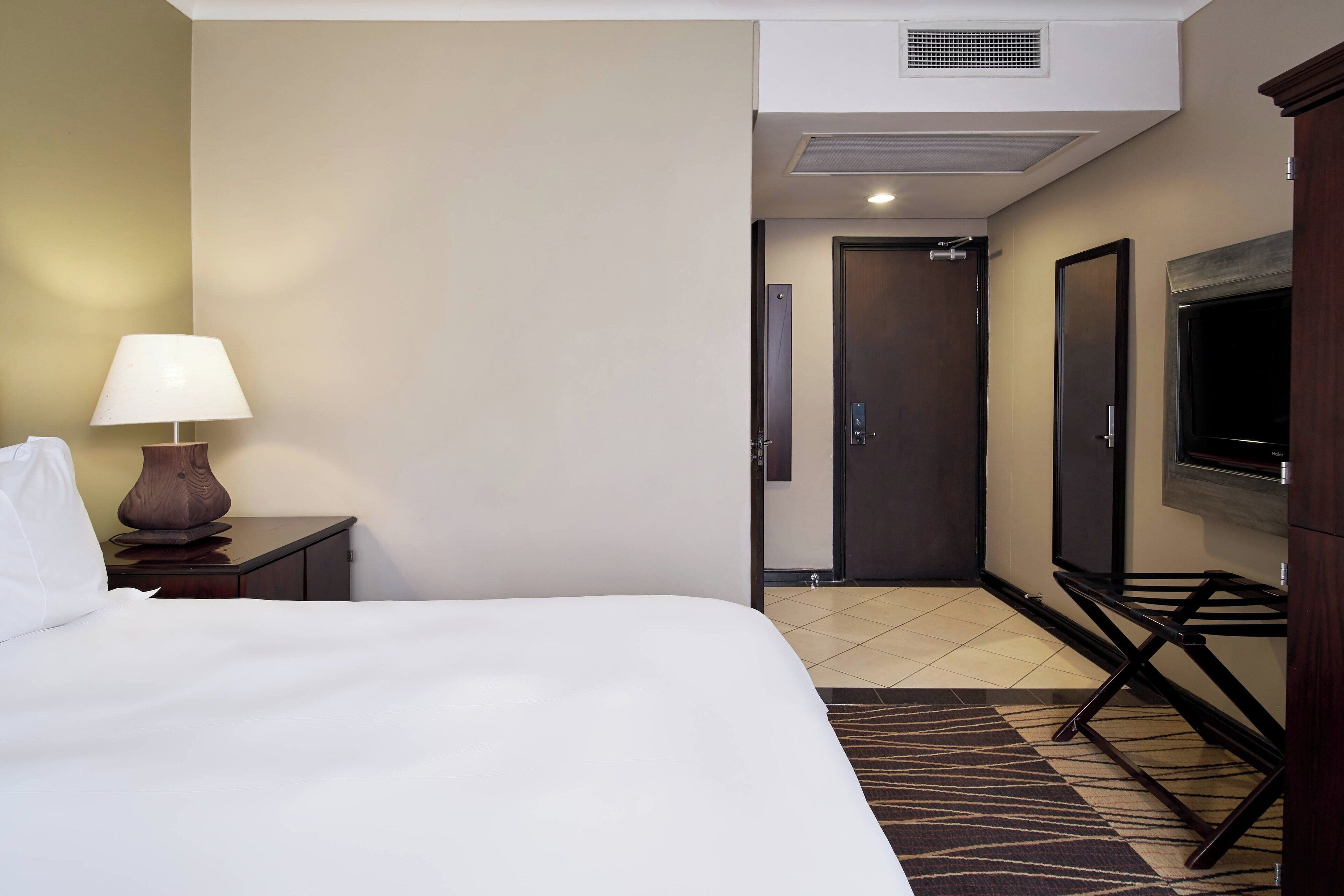 A hotel room with a bed, a nightstand with a lamp, a TV, and a door leading to a hallway.