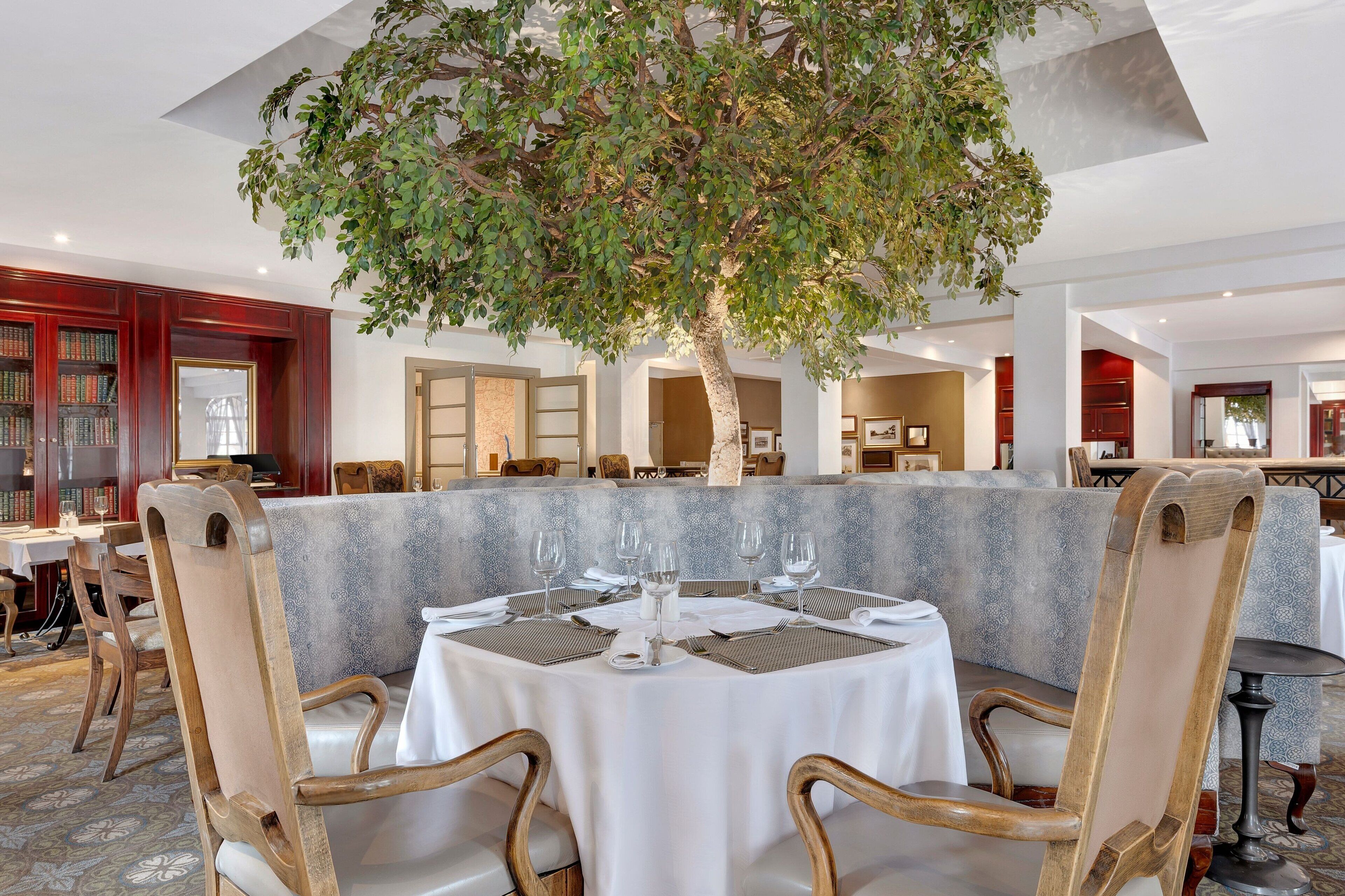 Een eethoek met een ronde tafel voor zes personen, omringd door houten stoelen. Een grote boom bevindt zich boven de tafel en zorgt voor schaduw. De ruimte heeft een combinatie van houten en stenen elementen, met een vloer met een patroon.